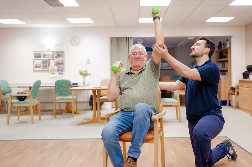 A South Coast physiotherapist supporting a client raising a dumbbell in his living room